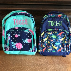 May include: Two personalized backpacks, one blue with a dinosaur print and the name "Tucker" embroidered in green, and the other teal with a floral print and the name "Tessa" embroidered in pink.