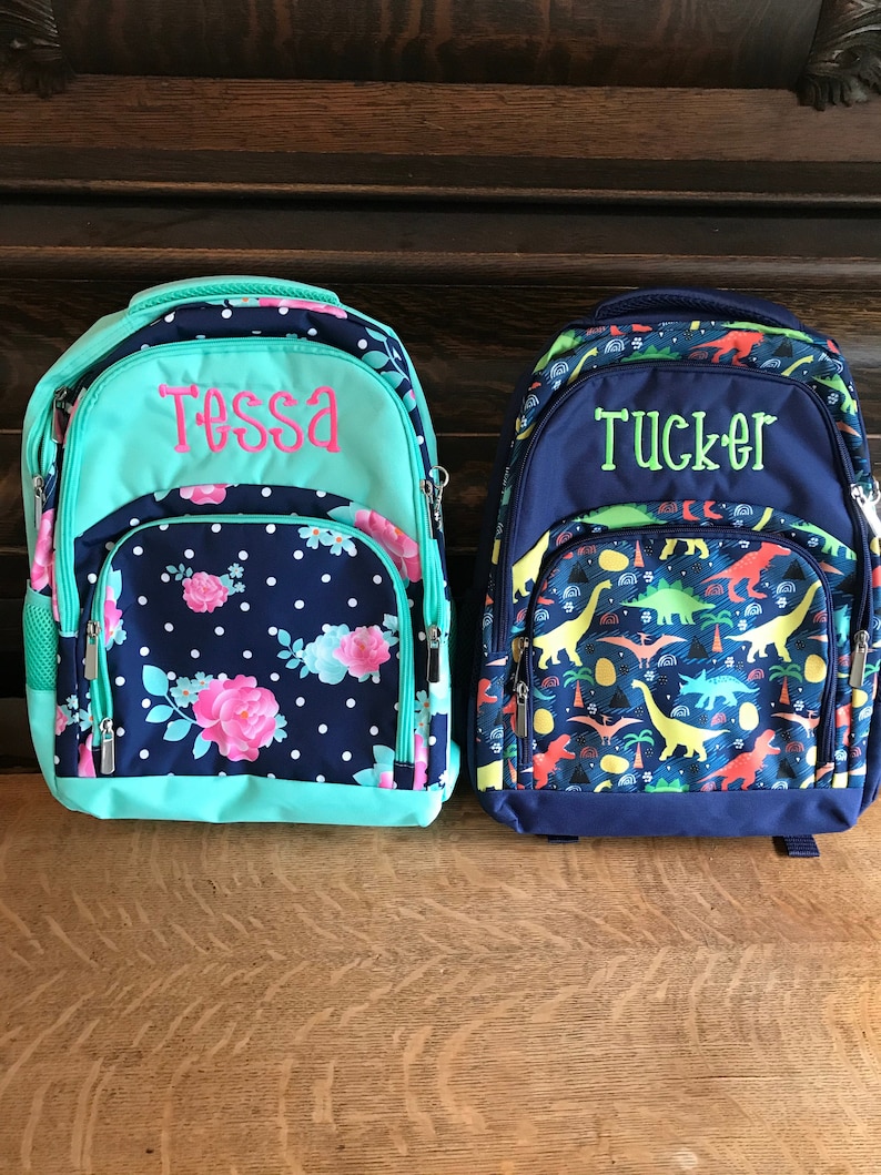 May include: Two personalized backpacks, one blue with a dinosaur print and the name "Tucker" embroidered in green, and the other teal with a floral print and the name "Tessa" embroidered in pink.