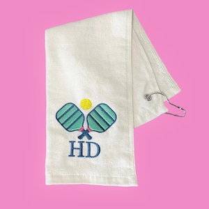 May include: White towel with embroidered pickleball paddles and ball in teal, blue, and yellow. The towel has the initials "HD" embroidered in blue. A silver metal clip is attached to the towel.