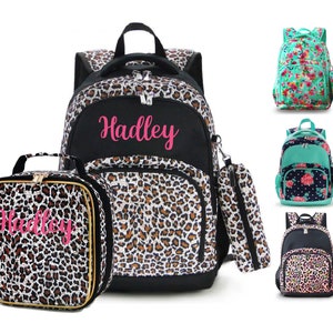 May include: A black and leopard print backpack and matching lunch bag set with the name "Hadley" in pink script. Additional backpacks in floral and leopard prints are also shown.