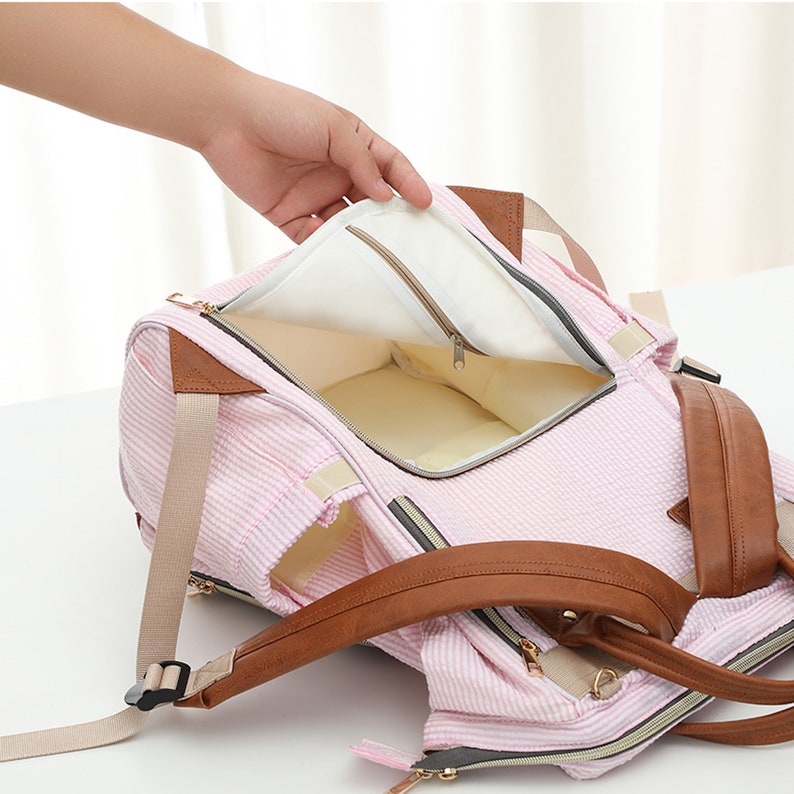 May include: Pink and white striped diaper bag with brown leather accents. The bag has multiple pockets and compartments, including a zippered pocket on the inside. The bag is open and the inside is lined with a light beige fabric.
