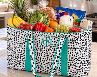 May include: A large rectangular tote bag with a black and white dalmatian-style spotted pattern and teal straps. The front features the embroidered monogram letters AFL in teal. The bag is filled with various groceries including produce, bread, and milk.