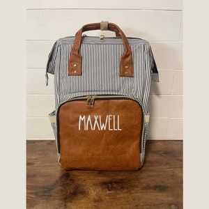 May include: A blue and white striped diaper bag with a brown faux leather front panel. The front panel has the name "MAXWELL" embroidered in white.