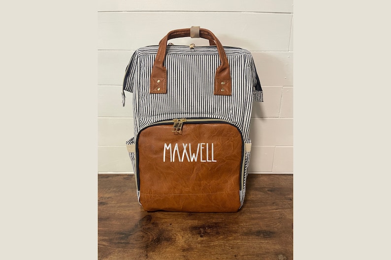 May include: A blue and white striped diaper bag with a brown faux leather front panel. The front panel has the name "MAXWELL" embroidered in white.