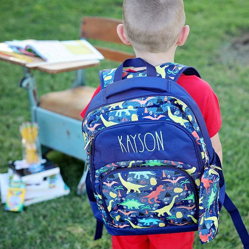 May include: A blue backpack with a dinosaur print and the name "Kayson" embroidered on the front. The backpack has a zippered pocket on the front and a mesh pocket on the side. The backpack is worn by a child wearing a red shirt.