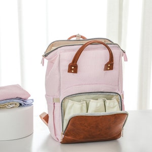 May include: A pink and white striped diaper bag backpack with a brown leather handle and zippered pockets. The bag has multiple interior pockets and a large main compartment.