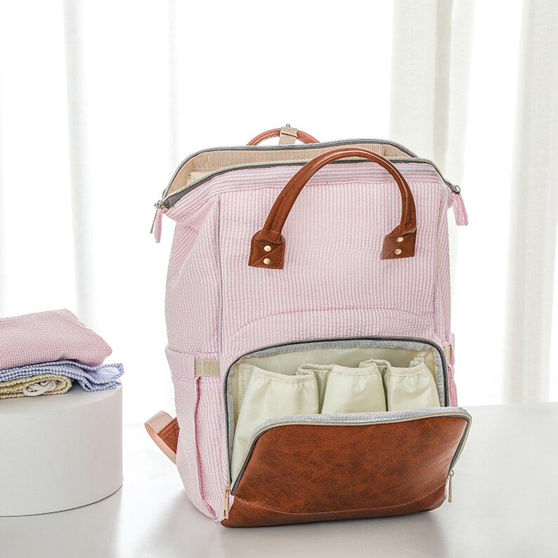 May include: A pink and white striped diaper bag backpack with a brown leather handle and zippered pockets. The bag has multiple interior pockets and a large main compartment.