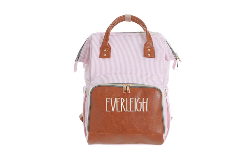 May include: A pink and white striped diaper bag with a brown faux leather bottom panel. The bag has a zippered pocket on the front with the word "EVERLEIGH" embroidered in white.