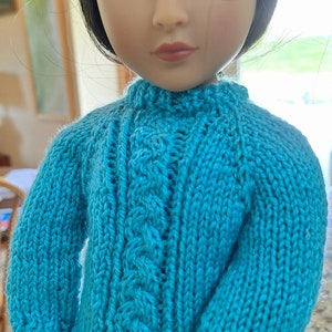May include: A turquoise knitted sweater with a cable knit pattern. The sweater is made for a doll and has long sleeves.