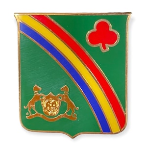 Pode incluir: Um broche de metal dourado com fundo verde e um arco-íris colorido. O broche apresenta um brasão dourado com dois cães e um escudo, e um trevo vermelho no canto superior direito.