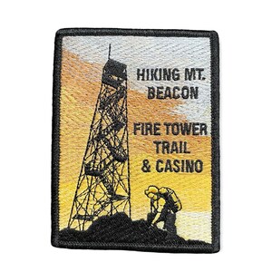 May include: An embroidered patch featuring a black silhouette of a fire tower on a yellow and orange sunset background. The text "Hiking Mt. Beacon Fire Tower Trail & Casino" is embroidered in black.