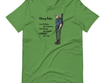Op de afbeelding: Groen T-shirt met een cartoonillustratie van een wandelaar en de tekst "Wandelregels: Laat niets achter behalve voetafdrukken, neem niets mee behalve foto's, dood niets behalve tijd."