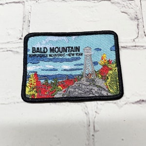 May include: A black and white embroidered patch with a blue background. The patch features a tower on a mountain with the text "Bald Mountain Adirondack Mountains - New York" in black.