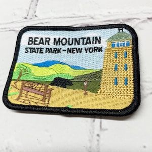 May include: Embroidered patch featuring a black bear, a wooden bench, a mountain range, and a tall tower with the text "Bear Mountain State Park - New York".