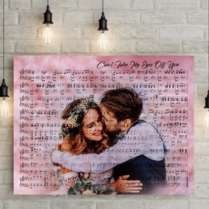 Op de afbeelding: Een canvas print met een omhelzend stel, over een bladmuziek met een roze aquarel achtergrond. De tekst "Can't Take My Eyes Off You" is in script geschreven. De print wordt verlicht door hangende Edison gloeilampen.