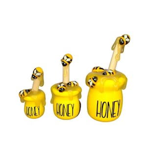 May include: Three yellow honey pot figurines with bees and a wooden honey dipper. The smallest pot is on the left, the middle pot is in the center, and the largest pot is on the right. Each pot has the word "Honey" written on the front.