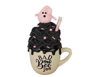 May include: A white ceramic mug with a black and pink ghost-shaped marshmallow topping, black whipped cream, pink sprinkles, and a pink and white striped straw. The mug has black text that reads "Bad and Boo jee".