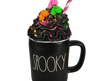 May include: Black ceramic mug with the word "SPOOKY" in white. The mug is topped with black whipped cream, colorful paint splatters, and three small skull decorations. A pink and white striped straw is inserted into the whipped cream.