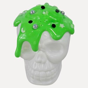 May include: A white skull figurine with bright green, drippy slime covering the top. The slime is decorated with small black circles, silver beads, and clear crystals. A white star and crescent moon are also visible.