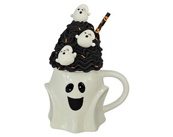 May include: A white ceramic Halloween mug shaped like a ghost with a smiling face. The mug is topped with black frosting and white ghost decorations. A straw with orange and black accents is in the frosting.