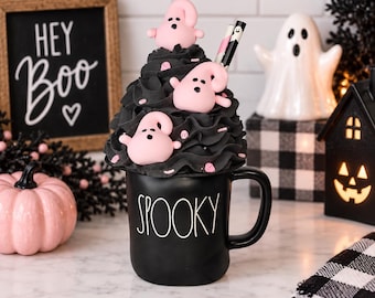 May include: A black ceramic mug with the word SPOOKY printed in white text. The mug is topped with a tall swirl of black faux whipped cream, decorated with three small light pink ghost figures and pink sprinkles, plus two patterned straws.