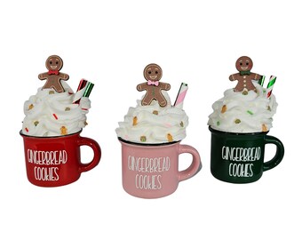 May include: Three decorative mugs with whipped cream, gingerbread men, and candy canes. The mugs are red, pink, and green, each with "GINGERBREAD COOKIES" in white. Festive holiday decor.