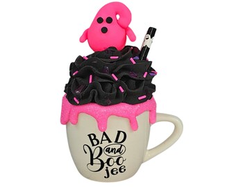 May include: A white ceramic mug with a pink and black frosting design. The mug has a pink rim and a pink drip design. The mug has the text "Bad and Boo jee" written on it in black.