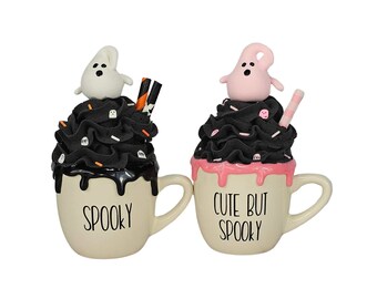 May include: Two ceramic mugs with black and white frosting and ghost toppers. One mug says "Spooky" and the other says "Cute But Spooky".