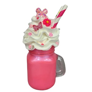 May include: A pink milkshake in a mason jar with a handle, topped with whipped cream, pink and white candies, and a pink and white striped straw. The whipped cream is decorated with a pink butterfly and a pink flower.
