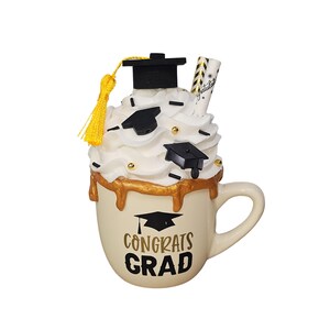May include: White ceramic mug with a gold drip design and the text "Congrats Grad" in gold lettering. The mug is topped with white frosting, black sprinkles, gold sprinkles, and black graduation caps.