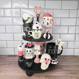 K&ouml;nnte beinhalten: Ein zweistufiges schwarzes Tablett, das mit Halloween-Artikeln dekoriert ist. Die obere Stufe zeigt eine schwarz-wei&szlig;e Kaffeetasse mit dem Text "Candy Cute", einen schwarz-wei&szlig;en Cupcake, einen schwarz-wei&szlig;en K&uuml;rbis und eine schwarz-wei&szlig;e Kaffeetasse mit dem Text "Boo". Die untere Stufe zeigt einen schwarz-wei&szlig;en Geist, einen schwarz-wei&szlig;en Sch&auml;del, eine schwarz-wei&szlig;e Kaffeetasse mit dem Text "Spooky", einen schwarz-wei&szlig;en Kessel, einen schwarz-wei&szlig;en Cupcake und einen schwarz-wei&szlig;en Geist.