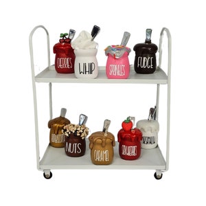 May include: White metal two-tiered serving cart with eight ceramic jars filled with different toppings for ice cream. The jars are labeled "Cherries", "Whip", "Sprinkles", "Fudge", "Butterscotch", "Nuts", "Caramel", "Strawberries", and "Marshmallow".