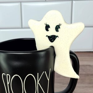 K&ouml;nnte beinhalten: Eine schwarze Keramiktasse mit dem Wort "Spooky" darauf. Ein wei&szlig;er Geisterkeks mit Glitzeraugen und einem L&auml;cheln sitzt auf dem Rand der Tasse.