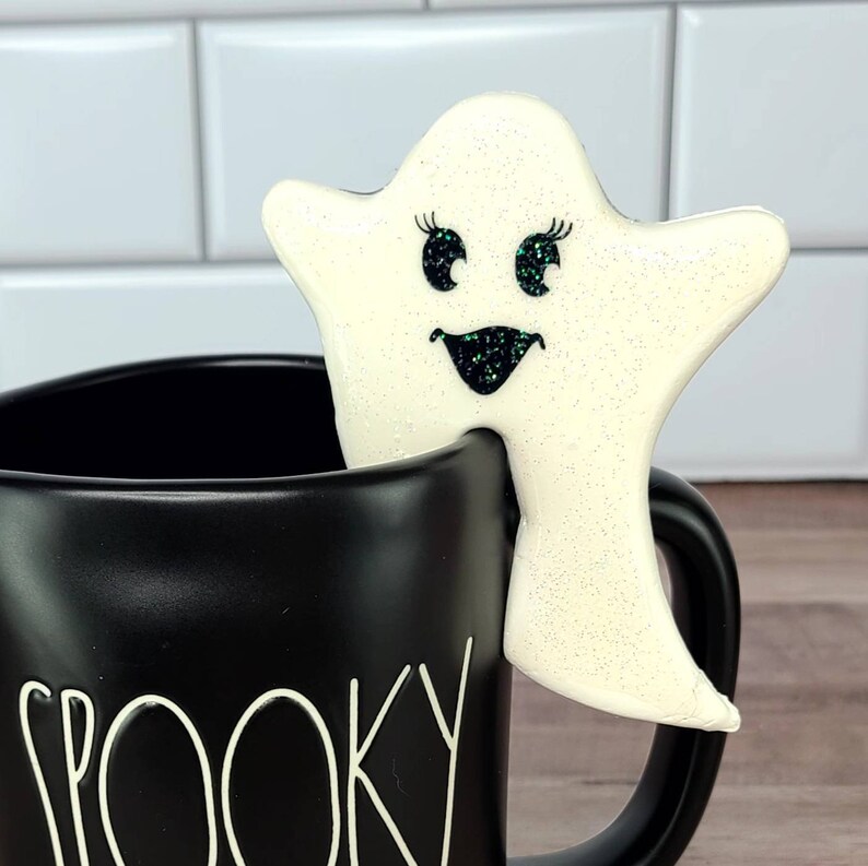 K&ouml;nnte beinhalten: Eine schwarze Keramiktasse mit dem Wort "Spooky" darauf. Ein wei&szlig;er Geisterkeks mit Glitzeraugen und einem L&auml;cheln sitzt auf dem Rand der Tasse.
