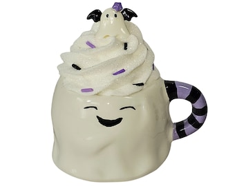May include: A white ceramic Halloween-themed mug shaped like a ghost with a smiling face. The mug is topped with a swirl of white whipped cream with purple and black sprinkles. A small ghost with bat wings sits on top. The handle has black and purple stripes.