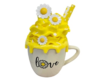 May include: A white ceramic mug with a yellow glaze dripping down the sides. The mug has a black "love" inscription with a daisy graphic. The mug is topped with yellow whipped cream and white daisies.