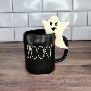 K&ouml;nnte beinhalten: Schwarze Keramiktasse mit dem Wort "Spooky" in wei&szlig; auf der Vorderseite. Ein wei&szlig;er Geisterkeks mit schwarzen Augen und einem L&auml;cheln sitzt auf dem Rand der Tasse.