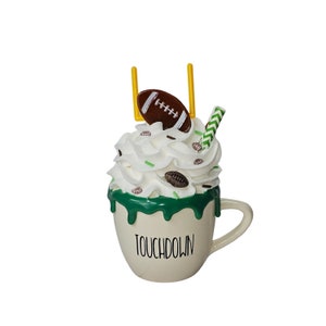 May include: A decorative mug with a football theme. The mug is white with the word "TOUCHDOWN" printed on it. It is topped with whipped cream, football-shaped sprinkles, a football, a goalpost, and a green and white striped straw.