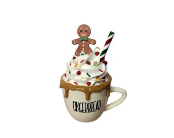 May include: White ceramic mug with a gingerbread man on top, topped with whipped cream, sprinkles, and a candy cane straw. The mug says "Gingerbread" on the side.