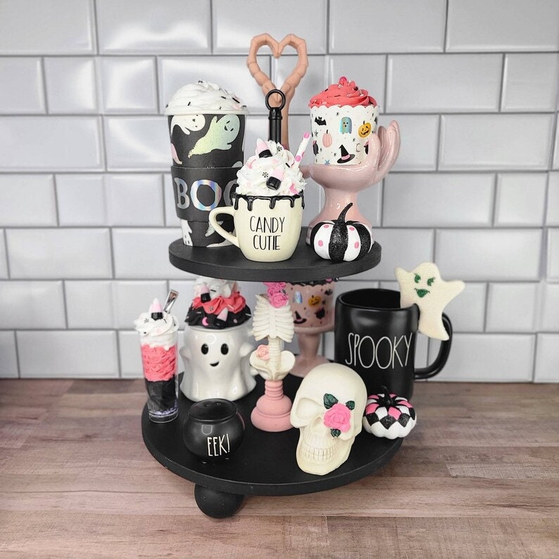 K&ouml;nnte beinhalten: Ein zweistufiges schwarzes Tablett, das mit Halloween-Artikeln dekoriert ist. Die obere Ebene zeigt einen schwarz-wei&szlig;en K&uuml;rbis, einen rosa Cupcake mit einem schwarz-wei&szlig;en Muster, eine schwarz-wei&szlig;e Kaffeetasse mit dem Text "Candy Cutie" und eine schwarz-wei&szlig;e Kaffeetasse mit einem Geistermotiv. Die untere Ebene zeigt einen schwarz-wei&szlig;en Geist, ein schwarz-wei&szlig;es Skelett, eine schwarze Tasse mit dem Text "Spooky", einen schwarzen Kessel mit dem Text "Eeek", einen rosa-wei&szlig;en Cupcake, einen wei&szlig;en Sch&auml;del mit einer rosa Rose und einen schwarz-wei&szlig;en K&uuml;rbis.