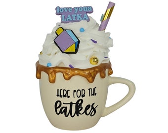 May include: A white ceramic mug with a gold rim and a brown glaze dripping down the side. The mug has a white whipped cream topping with sprinkles and a purple and yellow dreidel on top. The mug says "Here for the latkes" and has a pink sticker that says "Love youa latka" on the side.