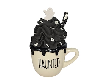 May include: A white ceramic mug with black dripping icing and black whipped cream topped with white ghost sprinkles and a black and white striped straw. The mug says "HAUNTED" in black lettering.