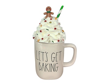 May include: A cream-colored mug with the text "LET'S GET BAKING" in black lettering. The mug is topped with a swirl of white whipped cream, decorated with sprinkles and a gingerbread man. A green and white striped straw is inserted into the whipped cream.