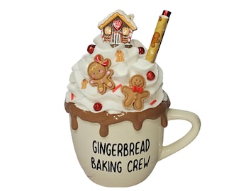 May include: White ceramic mug with a brown chocolate glaze and whipped cream topping. The mug has a gingerbread house, gingerbread people, and red sprinkles on top. The mug has the text "Gingerbread Baking Crew" on the front.