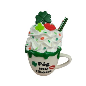 May include: White ceramic mug with green and white whipped cream, green shamrocks, and a red lip design. The mug has green icing and says "Póg mo thóin" in black lettering.