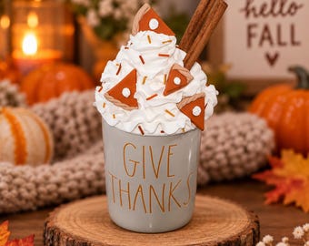 May include: A light gray ceramic mug with the text GIVE THANKS in orange lettering. The mug is topped with white faux whipped cream, orange pumpkin pie slice decorations, and two brown cinnamon stick accents.
