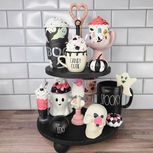K&ouml;nnte beinhalten: Ein dreistufiges Tablett, das mit Halloween-Artikeln dekoriert ist. Das Tablett zeigt eine schwarz-wei&szlig;e Geister-Tasse, eine schwarze Tasse mit dem Wort "Spooky" darauf, einen Sch&auml;del mit einer rosa Rose, eine Geisterfigur, einen schwarz-wei&szlig;en K&uuml;rbis und einen rosa-wei&szlig;en Cupcake.