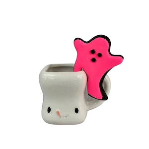 May include: A white ceramic mug with a smiling face and a pink ghost figurine inside. The mug has a handle and a textured surface.
