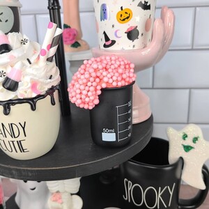 K&ouml;nnte beinhalten: Ein dreistufiges Tablett, das f&uuml;r Halloween dekoriert ist, mit einer schwarz-wei&szlig;en Tasse, auf der "Spooky" steht, einer wei&szlig;en Tasse mit schwarzer Aufschrift "Candy Cutie", einer rosa Hand, die eine wei&szlig;e Tasse mit einem Halloween-Muster h&auml;lt, und einem schwarzen Becher mit rosa Schleim darauf.