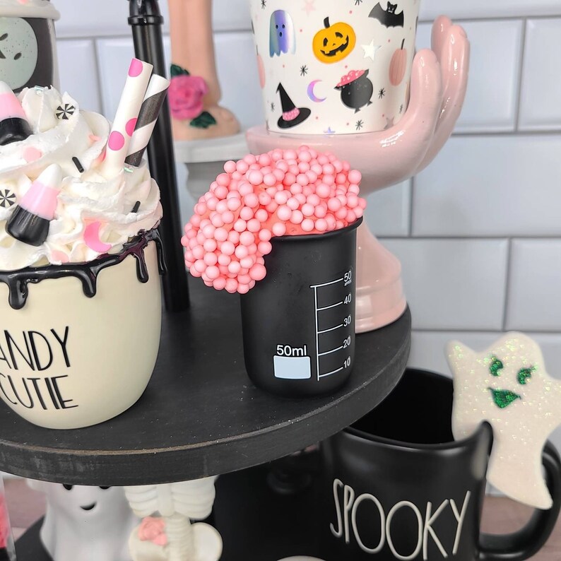 K&ouml;nnte beinhalten: Ein dreistufiges Tablett, das f&uuml;r Halloween dekoriert ist, mit einer schwarz-wei&szlig;en Tasse, auf der "Spooky" steht, einer wei&szlig;en Tasse mit schwarzer Aufschrift "Candy Cutie", einer rosa Hand, die eine wei&szlig;e Tasse mit einem Halloween-Muster h&auml;lt, und einem schwarzen Becher mit rosa Schleim darauf.